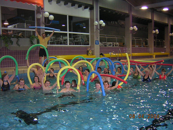 Eine Gruppe von Menschen schwimmt im Hallenbad und hält bunte Poolnudeln hoch.