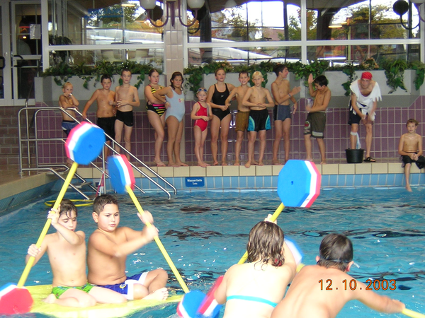 Kinder spielen mit bunten Paddeln im Wasser des Hallenbads, während eine Gruppe am Beckenrand zuschaut.