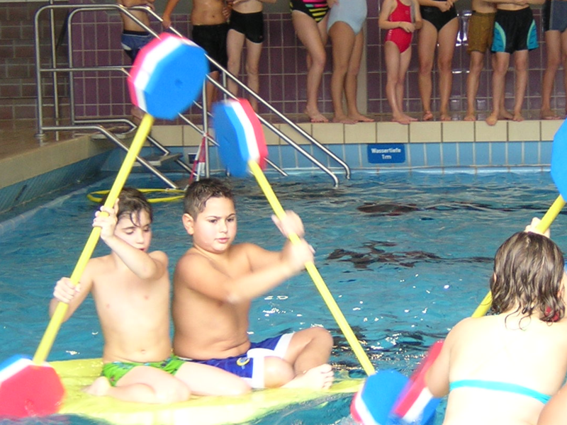 Kinder spielen mit bunten Paddeln im Wasser des Hallenbads, während eine Gruppe am Beckenrand zuschaut.