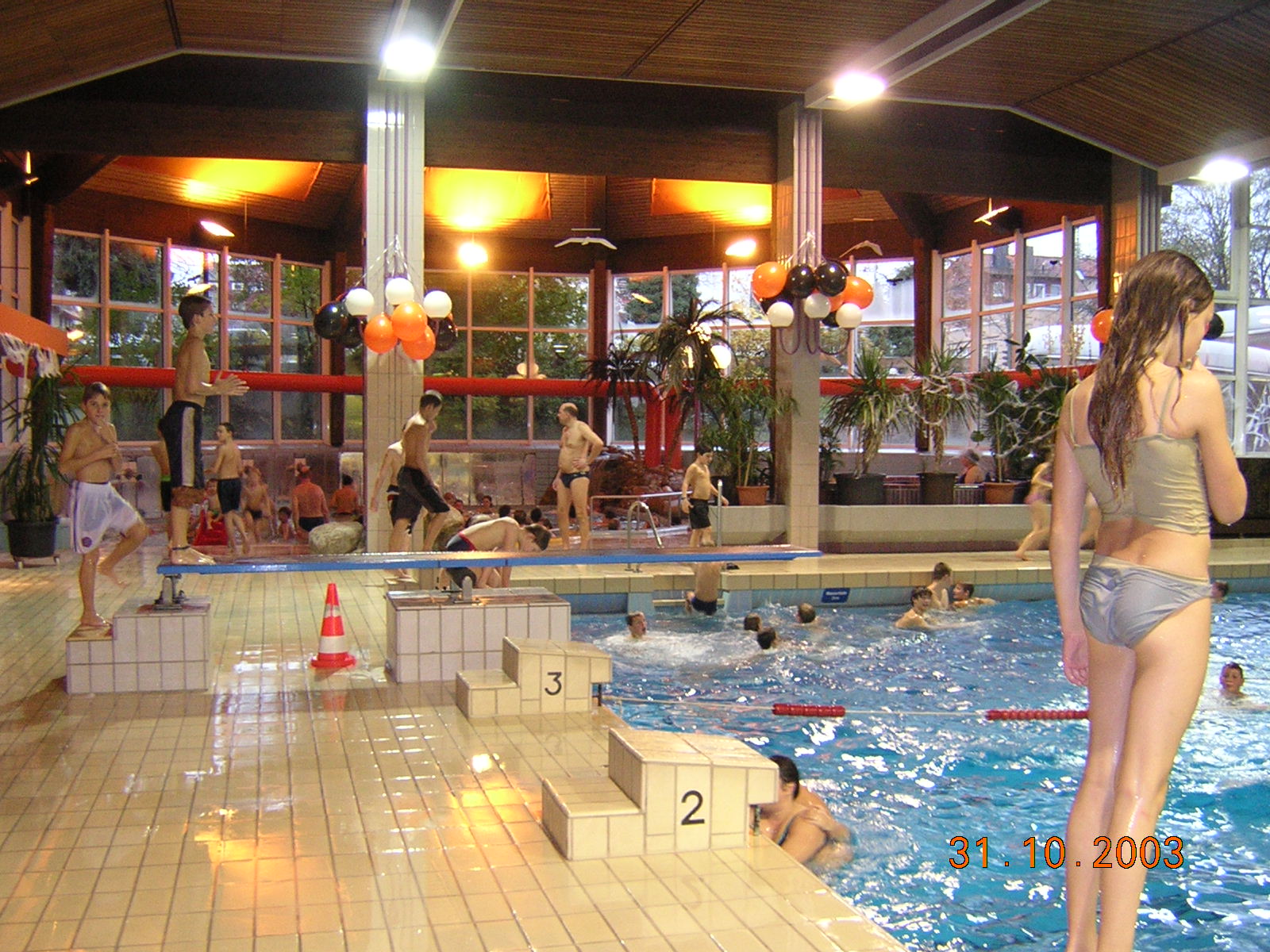 In der gut beleuchteten Schwimmhalle mit großer Fensterfront sind zahlreiche Menschen im und am Wasser sowie bunte Luftballons dekoriert.