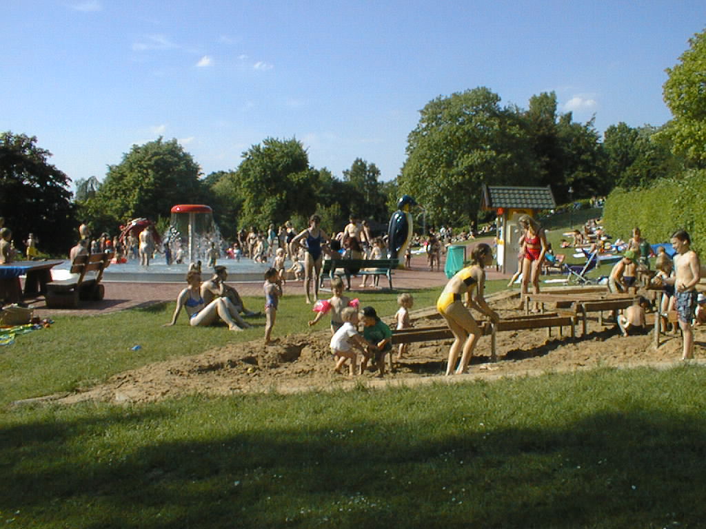 Im Bergbad  spielen Kinder in einem Sandbereich, während andere im Hintergrund in einem Wasserbereich planschen, umgeben von Bäumen und Wiesen.