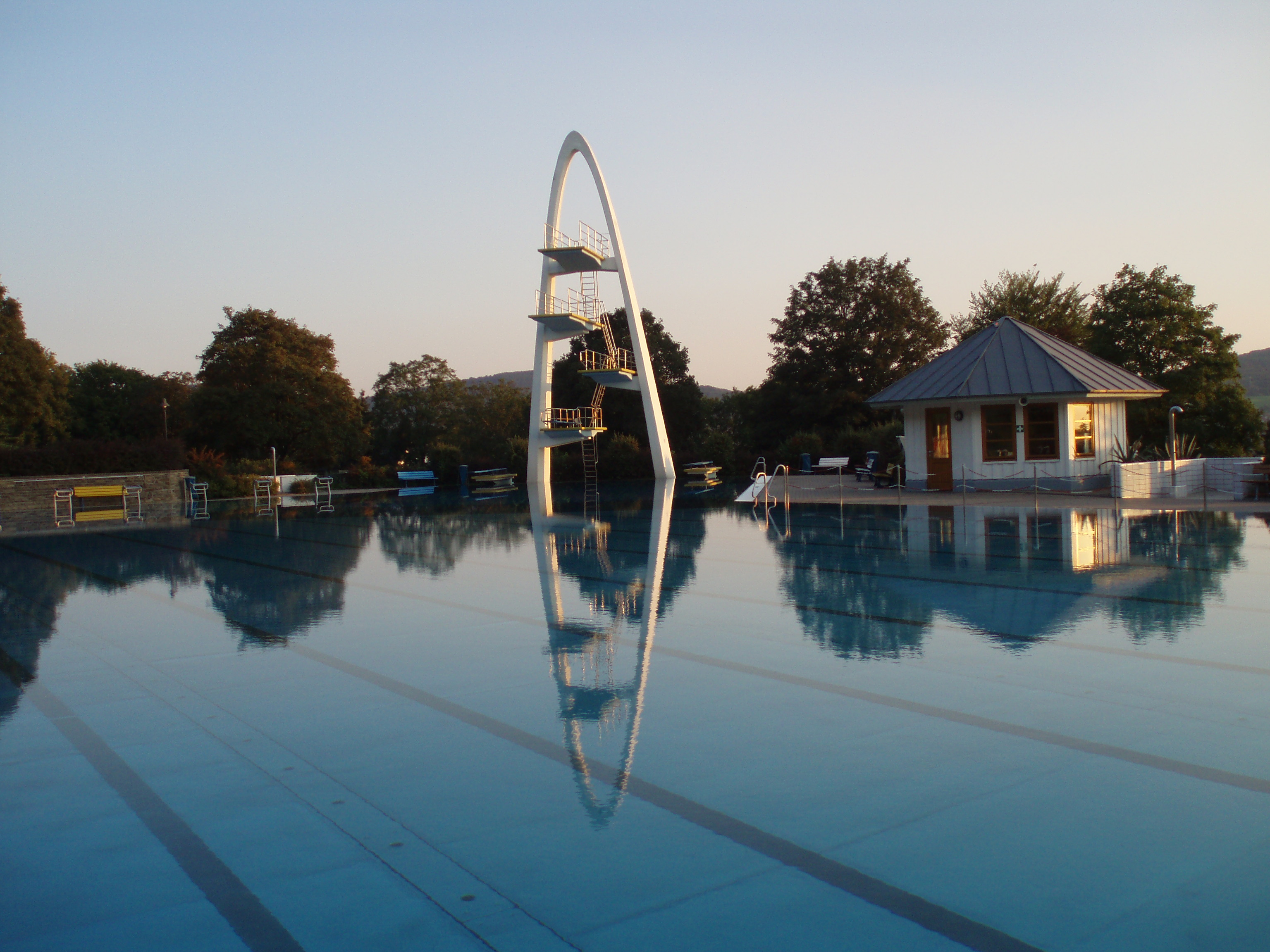 Ein großes, ruhiges Schwimmbecken mit einem markanten, modernen Sprungturm und einem kleinen Gebäude daneben, umgeben von Bäumen in der Abenddämmerung.